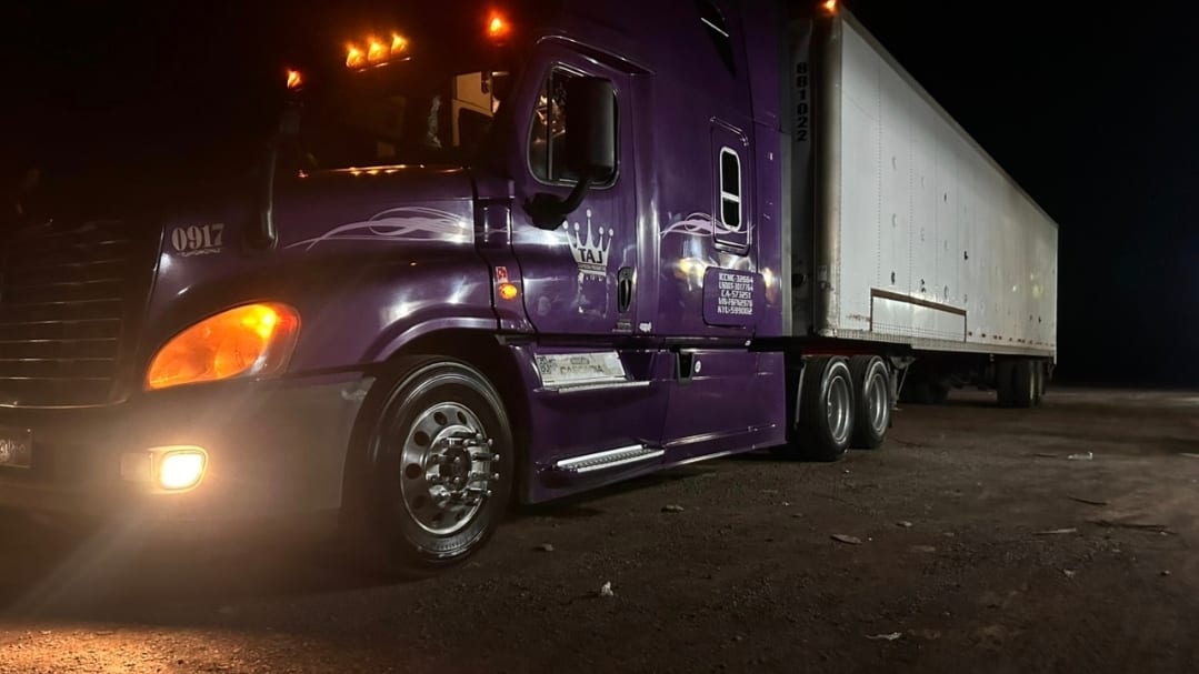 Tractocamión morado de Romo's Transportes con caja seca durante una operación nocturna, con las luces encendidas.
